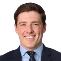 A smiling man with short brown hair wearing a dark suit, light blue shirt, and patterned tie, posed against a white background.