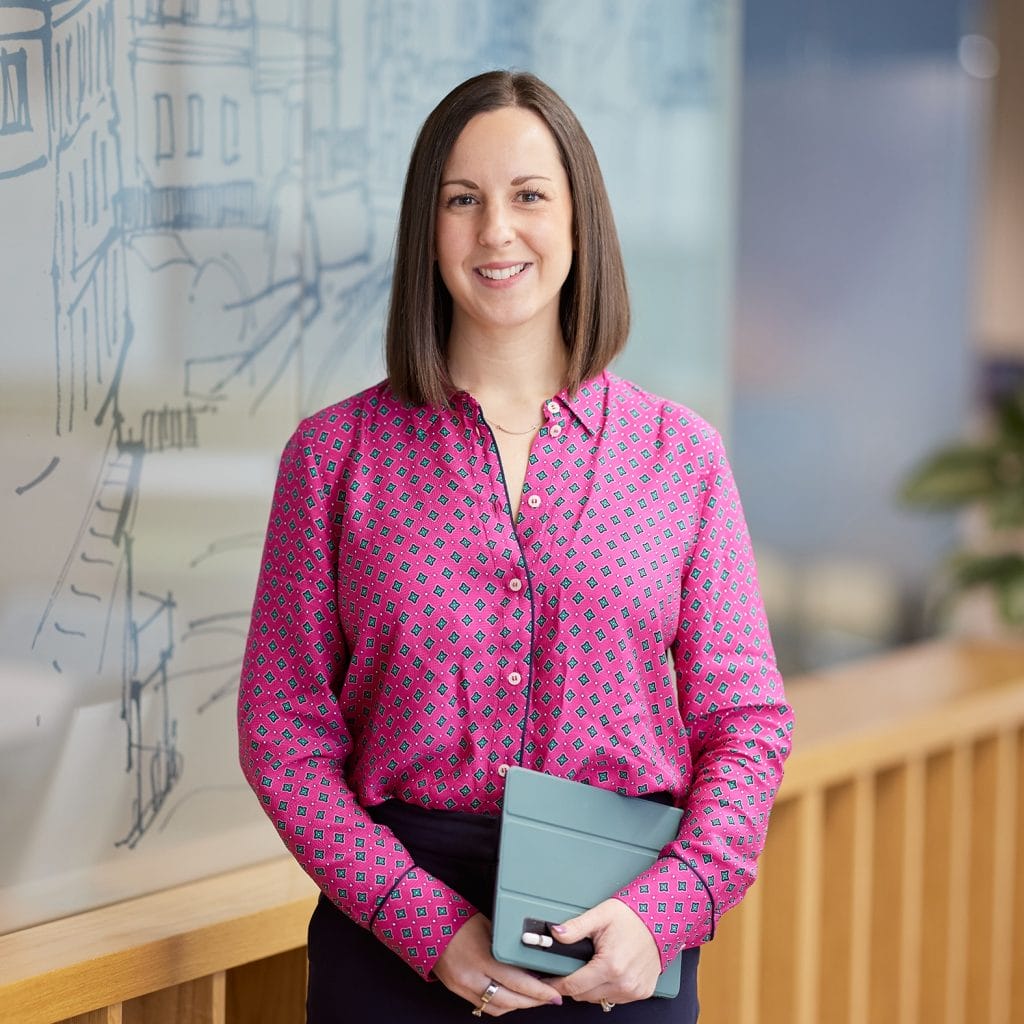 A woman in a pink patterned blouse stands indoors holding a closed tablet. She is smiling and standing near a glass wall with a drawing on it.