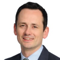 Portrait of a person in a suit and tie, smiling, with short dark hair. White background.