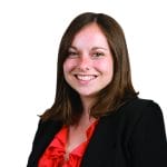A smiling person with shoulder-length brown hair wearing a black blazer and red blouse against a white background.