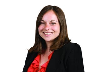 A smiling person with shoulder-length brown hair wearing a black blazer and red blouse against a white background.