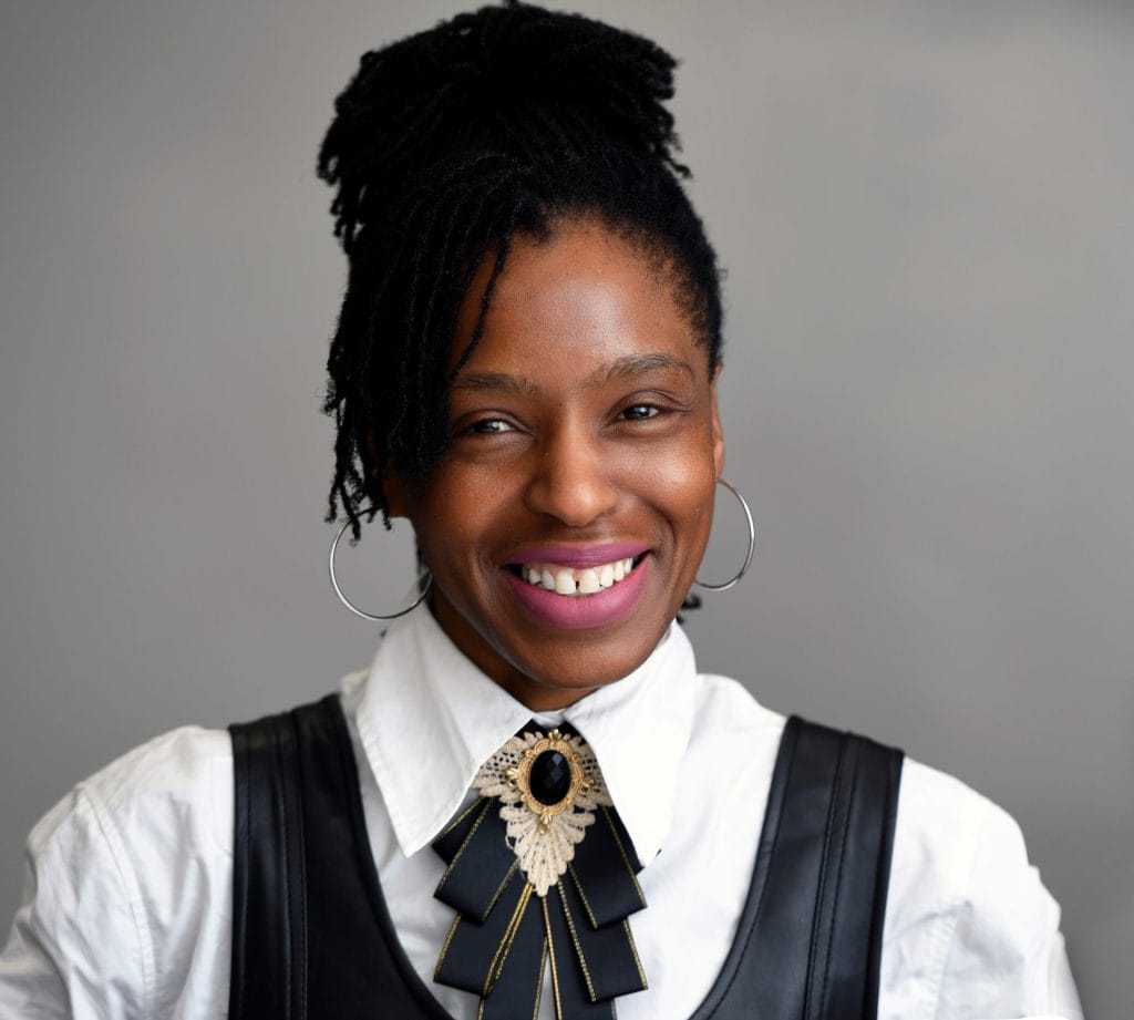 A black woman with dreadlocks wearing a vest and tie.