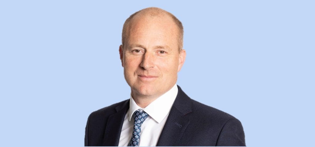 A man in a suit and tie is standing in front of a blue background.