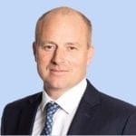 A man in a suit and tie is standing in front of a blue background.