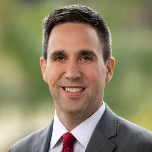 Professional headshot of a smiling man wearing a gray suit and red tie with a blurred natural background.