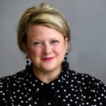 A smiling woman with short blonde hair, wearing black and white polka dot blouse and black earrings, poses against a gray background.