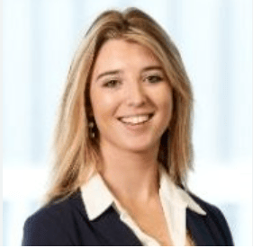 Professional headshot of a smiling woman with shoulder-length blonde hair, wearing a navy blazer and white blouse, against a light blue background.