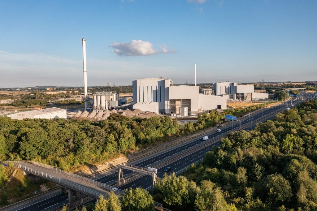 Aerial view of an industrial complex with multiple buildings and chimneys next to a highway surrounded by trees and greenery under clear blue sky.