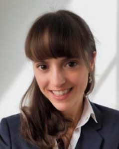 A woman with long brown hair and bangs, wearing a dark blazer and white shirt, smiling in front of a light background.