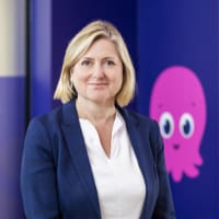 A person in a blue blazer and white shirt stands in front of a navy blue wall with a pink cartoon octopus graphic.