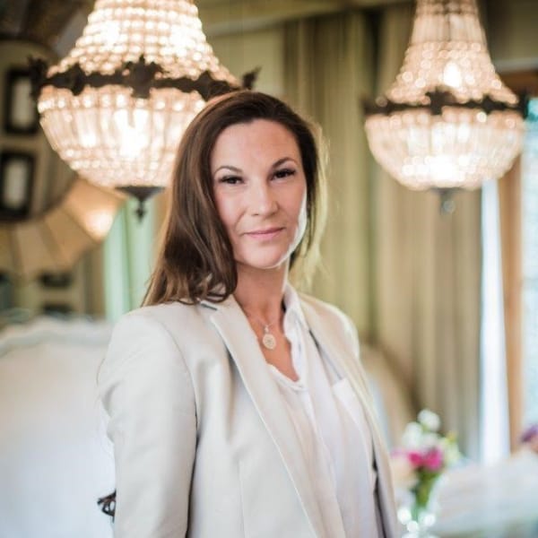 A person with long hair is standing indoors. They are wearing a light-colored blazer and shirt, with elegant chandeliers and a blurred background.