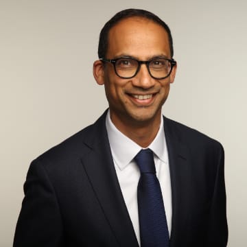 A man wearing glasses and a dark suit with a white shirt and blue tie smiles at the camera against a plain light background.