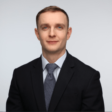 A man in a dark suit, light blue shirt, and patterned tie poses for a professional headshot against a plain light background.