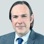 A man with straight brown hair, wearing a gray suit, white shirt, and blue patterned tie, smiles slightly against a plain light gray background.