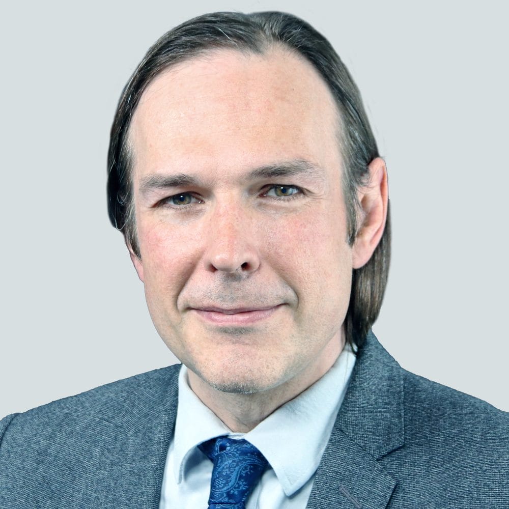 A man with straight brown hair, wearing a gray suit, white shirt, and blue patterned tie, smiles slightly against a plain light gray background.